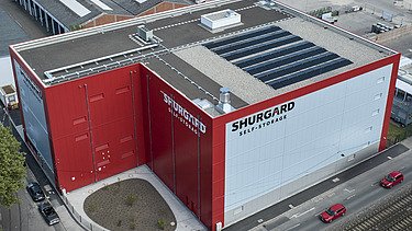 Luftaufnahme eines modernen Selfstorage-Gebäudes mit roter und weißer Fassade. Auf dem Flachdach sind Solarpaneele installiert, umgeben von Straßen und Industriegebäuden.