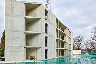 Baustelle mit Betongebäuden, Stahlträgern und Bauarbeitern mit Sicherheitsausrüstung unter teils bewölktem Himmel