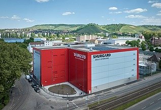 Großes Selfstorage-Gebäude mit roter und weißer Fassade, gelegen an einer Straße mit Straßenbahnschienen. Im Hintergrund grüne Hügel, Wohnhäuser und blauer Himmel mit wenigen Wolken.