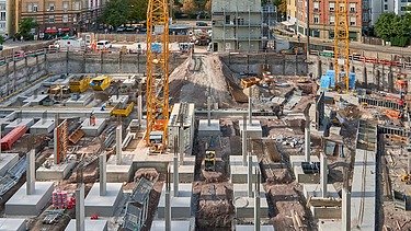 Große Baustelle mit gelben Baukränen, Betonfundamenten und zahlreichen Stützen, umgeben von mehrstöckigen Wohngebäuden. Im Hintergrund sind Bäume und ein teilweise bewölkter Himmel sichtbar.