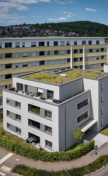 Luftaufnahme einer zeitgemäßen Wohnanlage mit mehreren Gebäuden, hellen Fassaden und begrünten Flachdächern. Die Anlage liegt an einer Straßenecke, umgeben von Gehwegen und Grünflächen, mit Blick auf eine hügelige Landschaft im Hintergrund.