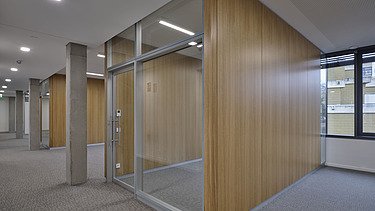 Innenansicht eines Bürobereichs mit Glaswand und Holzpaneelen, kombiniert mit Betonstützen und Teppichboden. Der Raum ist hell beleuchtet und verfügt über große Fenster für Tageslicht.