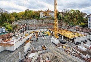 Große Baustelle mit gelbem Baukran, Betonfundamenten und Schalungen, umgeben von Baugeräten und Materialien. Im Hintergrund eine Felswand, Bäume und Wohnhäuser unter bewölktem Himmel.
