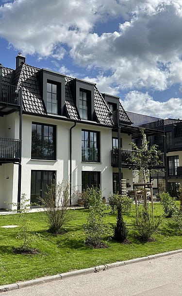 Mehrfamilienhäuser mit dunklen Dächern, großen Balkonen und grüner Gartenanlage unter blauem Himmel mit Wolken.