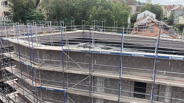 Mehrstöckiger Rohbau mit umlaufendem Baugerüst, dunkler Betonfassade und offenem Dachbereich, umgeben von Bäumen und angrenzenden Wohnhäusern.