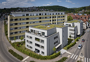 Luftaufnahme einer zeitgemäßen Wohnanlage mit mehreren Gebäuden, hellen Fassaden und begrünten Flachdächern. Die Anlage liegt an einer Straßenecke, umgeben von Gehwegen und Grünflächen, mit Blick auf eine hügelige Landschaft im Hintergrund.