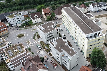 Luftaufnahme eines Wohngebiets mit mehreren modernen Mehrfamilienhäusern, einem großen gelben Wohnblock und kleineren Gebäuden rund um einen Kreisverkehr. Im Hintergrund sind Grünflächen und eine Straße sichtbar.