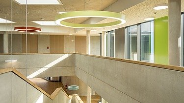 Innenansicht eines offenen Atriums mit Sichtbetonwänden, großen Oberlichtern und runden, farbigen Deckenleuchten. Die Architektur kombiniert klare Linien mit Holzdetails an den Geländern und viel natürlichem Licht durch große Fensterflächen.