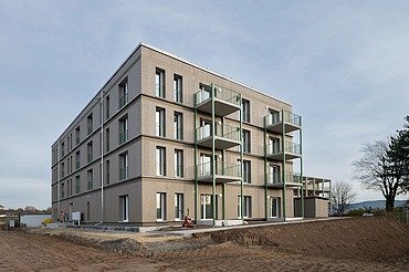 Außenansicht eines viergeschossigen Wohngebäudes mit klarer, moderner Fassadengestaltung und mehreren Balkonen. Das Gebäude steht noch auf einer unbefestigten Baustellenfläche, im Hintergrund Bäume und offener Himmel.