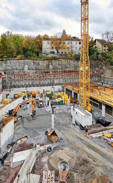 Große Baustelle mit gelbem Baukran, Betonfundamenten und Schalungen, umgeben von Baugeräten und Materialien. Im Hintergrund eine Felswand, Bäume und Wohnhäuser unter bewölktem Himmel.