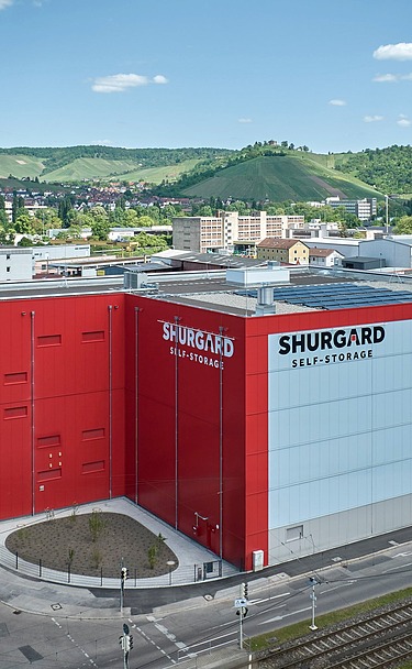 Großes Selfstorage-Gebäude mit roter und weißer Fassade, gelegen an einer Straße mit Straßenbahnschienen. Im Hintergrund grüne Hügel, Wohnhäuser und blauer Himmel mit wenigen Wolken.