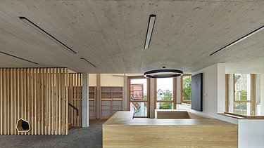 Innenansicht eines modernen Gebäudes mit Sichtbetondecke, Holzverkleidungen und offenen Regalflächen. Im Vordergrund steht eine helle, umlaufende Holzeinrichtung, dahinter verlaufen Fensterfronten, die Tageslicht in den Raum lassen. Eine offene Holztreppe mit vertikalen Lamellen prägt den Eingangs- und Aufenthaltsbereich.