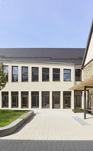 Innenhof eines modernen Gebäudekomplexes mit heller Putzfassade, großen Fensterfronten und einer gepflegten Grünfläche. Rechts befindet sich eine historische Natursteinwand mit Holzpergola, links ein kleiner Baum und Sitzbereiche. Der Hof ist offen gestaltet und mit hellen Pflastersteinen ausgelegt.