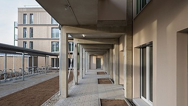 Blick in einen überdachten Laubengang eines modernen Wohngebäudes mit Betonstützen, hellen Fassaden und bodentiefen Fenstern. Rechts verläuft der Zugang zu den Wohnungen, links befinden sich überdachte Fahrradstellplätze und eine vorbereitete Außenfläche. Im Hintergrund ist die Holzfassade des Haupthauses sichtbar.