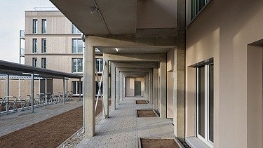 Blick in einen überdachten Laubengang eines modernen Wohngebäudes mit Betonstützen, hellen Fassaden und bodentiefen Fenstern. Rechts verläuft der Zugang zu den Wohnungen, links befinden sich überdachte Fahrradstellplätze und eine vorbereitete Außenfläche. Im Hintergrund ist die Holzfassade des Haupthauses sichtbar.