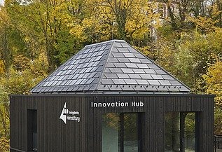 Außenansicht eines modernen Gebäudes mit dunkler Holzfassade und pyramidenförmigem Schieferdach, beschriftet mit „Innovation Hub“ und einem weiteren Logo. Das Gebäude steht inmitten herbstlicher Bäume und fügt sich in die bewaldete Umgebung ein.