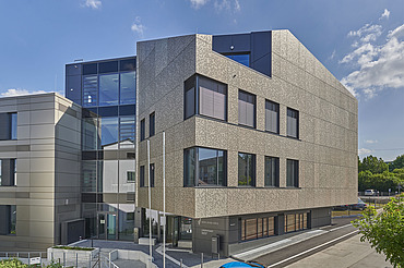 Mehrstöckiges Gebäude mit markanter, strukturierter Fassade in Beige und großen Glasflächen. Die Architektur kombiniert klare Linien mit modernen Materialien. Im Vordergrund ist eine Straße und ein Eingangsbereich sichtbar, im Hintergrund blauer Himmel mit wenigen Wolken.