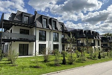 Mehrfamilienhäuser mit dunklen Dächern, großen Balkonen und grüner Gartenanlage unter blauem Himmel mit Wolken.