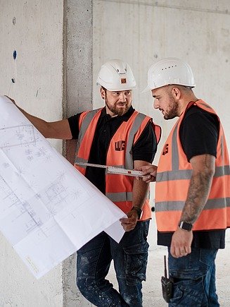 Zwei Poliere in Sicherheitswesten und Helmen prüfen Baupläne vor einer Betonwand.