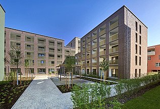 Mehrstöckige Wohnhäuser in moderner Architektur mit großen Balkonen und einem begrünten Innenhof, gepflasterten Wegen und jungen Bäumen unter klarem blauem Himmel.