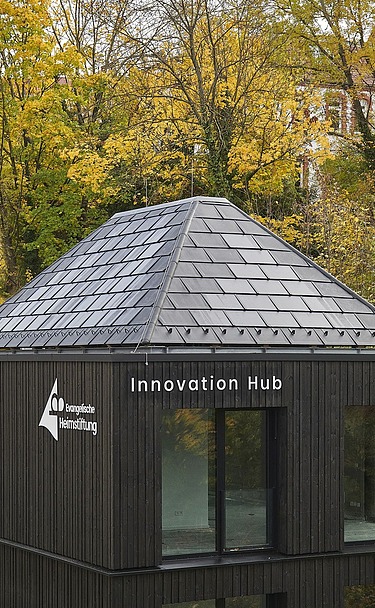 Außenansicht eines modernen Gebäudes mit dunkler Holzfassade und pyramidenförmigem Schieferdach, beschriftet mit „Innovation Hub“ und einem weiteren Logo. Das Gebäude steht inmitten herbstlicher Bäume und fügt sich in die bewaldete Umgebung ein.