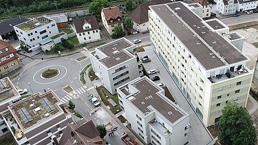 Drohnenaufnahme einer Wohnanlage mit mehreren mehrstöckigen Gebäuden in heller Fassade, angeordnet um einen Kreisverkehr. Die Umgebung umfasst Straßen, Einfamilienhäuser und Grünflächen, im Hintergrund ist eine Baumreihe und eine Bahntrasse sichtbar.