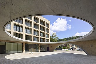 Ansicht eines mehrstöckigen Gebäudes durch eine große, ovale Öffnung in einer Betonüberdachung. Im Vordergrund ist ein gepflasterter Hof, im Hintergrund Bäume und blauer Himmel mit Wolken.