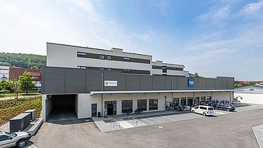 Modernes Wohn- und Geschäftshaus mit grauer und weißer Fassade. Im Erdgeschoss befindet sich ein Supermarkt mit sichtbarem Eingangsbereich und parkenden Autos davor. Im Hintergrund sind grüne Hügel und blauer Himmel zu sehen.