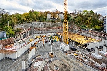 Große Baustelle mit gelbem Baukran, Betonfundamenten und Schalungen, umgeben von Baugeräten und Materialien. Im Hintergrund eine Felswand, Bäume und Wohnhäuser unter bewölktem Himmel.