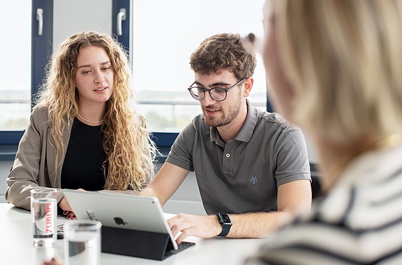 Studierende sitzen an einem Tisch in einem modernen Raum und arbeiten gemeinsam an einem Tablet