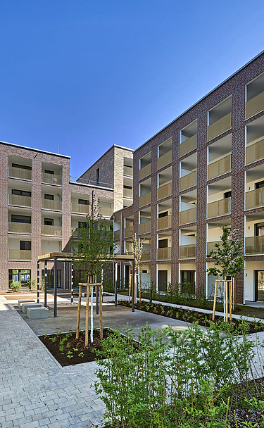 Mehrstöckige Wohnhäuser in moderner Architektur mit großen Balkonen und einem begrünten Innenhof, gepflasterten Wegen und jungen Bäumen unter klarem blauem Himmel.