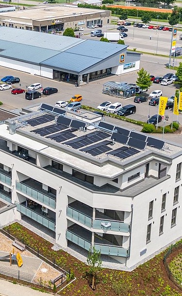 Luftaufnahme eines modernen Gebäudekomplexes mit heller Fassade und großen Balkonen. Auf den Flachdächern sind Solarpaneele installiert. Das Gebäude ist von Straßen, Grünflächen und weiteren Gewerbebauten umgeben.