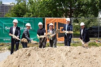 Baustelle mit Betongebäuden, Stahlträgern und Bauarbeitern mit Sicherheitsausrüstung unter teils bewölktem Himmel