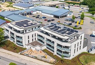 Luftaufnahme eines modernen Gebäudekomplexes mit heller Fassade und großen Balkonen. Auf den Flachdächern sind Solarpaneele installiert. Das Gebäude ist von Straßen, Grünflächen und weiteren Gewerbebauten umgeben.