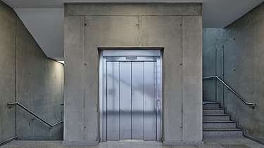 Ansicht eines schlichten, modernen Treppenhauses mit einem zentralen Edelstahl-Aufzug, flankiert von zwei Treppen aus Beton. Die Wände bestehen aus Sichtbeton, der Boden aus poliertem Stein, und die Beleuchtung erfolgt über eingelassene Deckenleuchten.