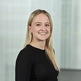 Ausbildungskoordinatorin mit langen, glatten Haaren in schwarzem Oberteil, vor einem hellen, strukturierten Hintergrund mit vertikalen Linien.