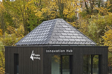 Außenansicht eines modernen Gebäudes mit dunkler Holzfassade und pyramidenförmigem Schieferdach, beschriftet mit „Innovation Hub“ und einem weiteren Logo. Das Gebäude steht inmitten herbstlicher Bäume und fügt sich in die bewaldete Umgebung ein.