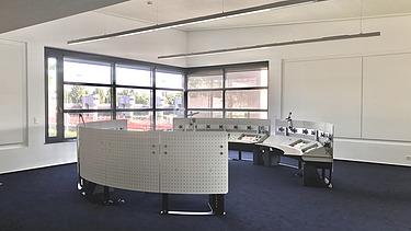 Heller Kontrollraum mit gebogenen Arbeitsstationen, großen Fenstern und moderner Beleuchtung.