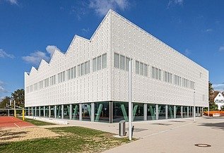 Außenansicht einer modernen Sporthalle mit heller Ziegelfassade, rhythmisch angeordneten Oberlichtern und grünen Stahlstützen im Erdgeschoss. Im Vordergrund befinden sich Sportflächen und Wege, umgeben von Bäumen und Schulgebäuden.