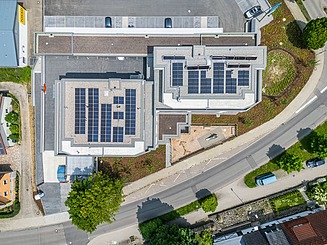 Luftaufnahme eines modernen Gebäudekomplexes mit großen Solarpanelen auf den Dächern, umgeben von Straßen, Grünflächen und angrenzenden Gewerbegebäuden.