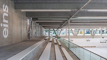 Blick auf die Tribüne einer Sporthalle mit Sitzstufen aus Sichtbeton und hellen Sitzflächen. Seitlich verlaufen große Fenster und tragende Stahlstützen, die Tageslicht in den Hallenraum lassen. An der Wand ist die Beschriftung „eins“ sichtbar.