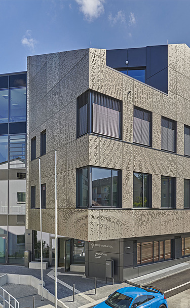 Mehrstöckiges Gebäude mit markanter, strukturierter Fassade in Beige und großen Glasflächen. Die Architektur kombiniert klare Linien mit modernen Materialien. Im Vordergrund ist eine Straße und ein Eingangsbereich sichtbar, im Hintergrund blauer Himmel mit wenigen Wolken.