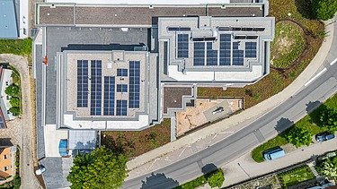 Draufsicht auf zwei moderne Gebäude mit Flachdächern, auf denen zahlreiche Solarpaneele installiert sind. Die Gebäude sind von Straßen, Grünflächen und weiteren Gewerbebauten umgeben.