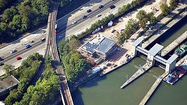 Baustelle an einem Wasserkanal neben Straßen und Bahntrassen, umgeben von Bäumen und Verkehr.