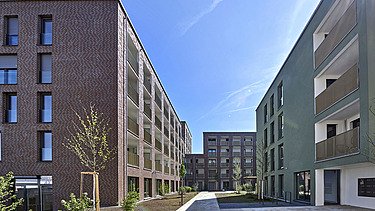 Mehrere moderne Mehrfamilienhäuser mit geradliniger Architektur, großen Balkonen und begrüntem Innenhof, unter klarem blauem Himmel.