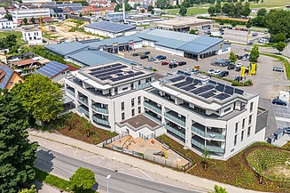 Luftaufnahme eines modernen Gebäudekomplexes mit großen Solarpanelen auf den Dächern, umgeben von Straßen, Grünflächen und angrenzenden Gewerbegebäuden.