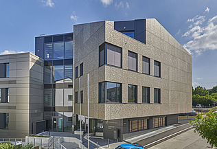 Mehrstöckiges Gebäude mit markanter, strukturierter Fassade in Beige und großen Glasflächen. Die Architektur kombiniert klare Linien mit modernen Materialien. Im Vordergrund ist eine Straße und ein Eingangsbereich sichtbar, im Hintergrund blauer Himmel mit wenigen Wolken.