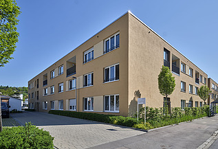 Ansicht eines modernen, dreigeschossigen Wohngebäudes mit hellgelber Fassade, großen Fenstern und Balkonen. Vor dem Gebäude befinden sich gepflegte Grünflächen und eine gepflasterte Zufahrt, im Hintergrund sind Bäume und blauer Himmel sichtbar.